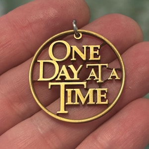 Peut inclure: Un pendentif doré avec les mots "ONE DAY AT A TIME" découpés dans un cadre circulaire. Le pendentif a une petite boucle en haut pour une chaîne. Le design est tenu dans la main d'une personne.