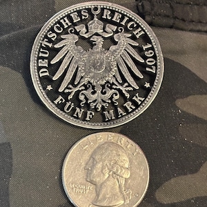 May include: A silver German 5 Mark coin from 1907, featuring an eagle emblem, next to a 1986 U.S. quarter. The coins are displayed on a camouflage fabric background. The German coin has the text "DEUTSCHES REICH" and "FÜNF MARK".