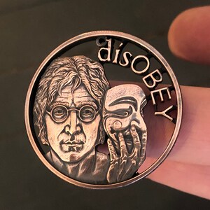 May include: A copper-coloured coin featuring a portrait of John Lennon and a mask with the word "disobey" inscribed around the edge.