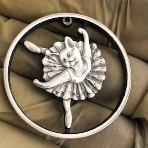 Op de afbeelding: Een zilverkleurige hanger met een gedetailleerd beeldhouwwerk van een kat die is verkleed als ballerina. De kat draagt een tutu en spitsschoenen en staat in een danspose. De hanger is rond en heeft bovenaan een gat om op te hangen.