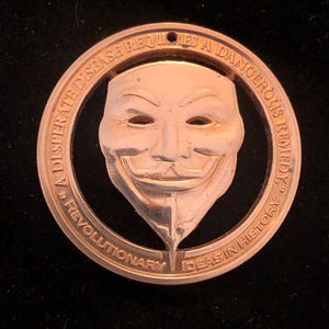 May include: A copper-colored round pendant with a cutout of the iconic V for Vendetta mask. The text around the mask reads "A desperate disease requires a dangerous remedy... Revolutionary ideas in history."