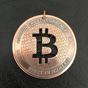 Puede incluir: Un colgante de criptomoneda Bitcoin de color cobre. El colgante redondo presenta el símbolo de Bitcoin recortado en el centro, rodeado de las palabras "Bitcoin Cryptocurrency" y "Vires in Numeris". El fondo tiene un patrón de código binario.