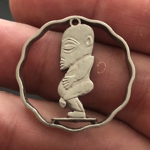 May include: A silver pendant featuring a detailed cutout of a person in a squatting position. The pendant is circular with a wavy edge and has a loop at the top for hanging.