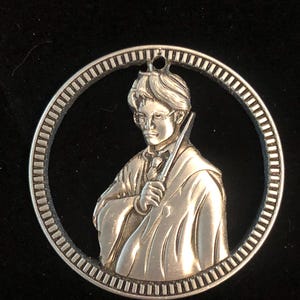 May include: A silver-colored circular pendant featuring a detailed relief of a figure holding a wand, framed by a textured border. The figure is wearing glasses and a robe. The pendant has a small hole at the top for hanging.