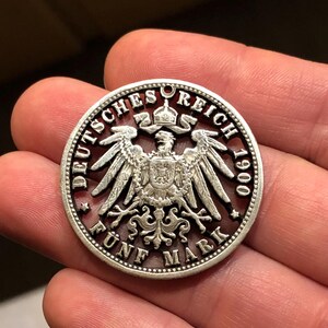 May include: A silver coin with the text "DEUTSCHES REICH 1900" and "FÜNF MARK" encircling a detailed eagle emblem. The coin has a decorative edge and a small hole at the top.