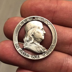 May include: A silver coin with a cutout design featuring a profile of Benjamin Franklin. The coin's outer ring includes the words "LIBERTY" and "IN GOD WE TRUST".