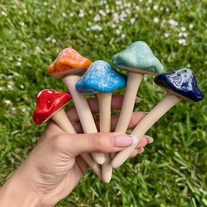 May include: A set of six ceramic mushroom garden stakes in various colors, including red, orange, blue, green, and white. The mushrooms have a smooth, glossy finish and are attached to wooden stakes.