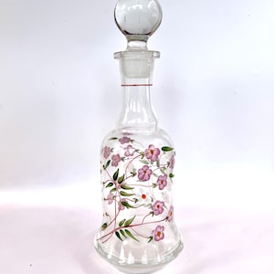 May include: A clear glass decanter with a round stopper. The decanter is decorated with pink flowers, green leaves, and red accents. The floral design wraps around the body of the bottle.
