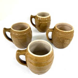 May include: Four small, brown ceramic mugs with handles. The mugs have a barrel-like shape with horizontal ridges and a light-colored interior. The mugs are arranged on a white surface.