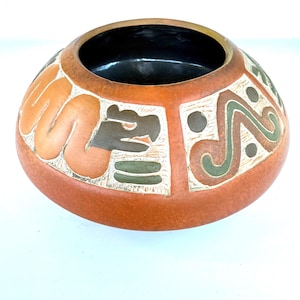 May include: A terracotta-colored ceramic bowl with a black interior. The exterior features carved designs in orange, white, and green, including geometric shapes and stylized animal figures. The bowl has a rounded shape and a wide opening.