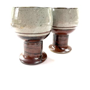 May include: Two handmade ceramic goblets. The bowls are light gray with dark speckles and a brown band. The stems are a dark brown with a textured appearance. The goblets are on a white background.