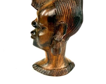 May include: A carved wooden sculpture of a human head in profile. The sculpture is a rich brown color with darker accents, and the wood grain is visible. The head has detailed features, including a defined nose, lips, and ear. The hair is styled with carved lines.