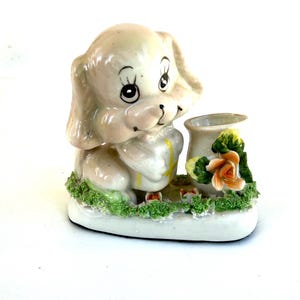 May include: A ceramic figurine of a puppy dog with large eyes and floppy ears. The puppy is holding a small vase with a flower arrangement. The base is decorated with green grass and small flowers. The figurine is painted in pastel colors.