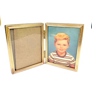 May include: A vintage gold-colored, hinged picture frame. One side holds a photograph of a young boy in a striped shirt. The other side is empty. The frame is open, revealing the photo and the backing.