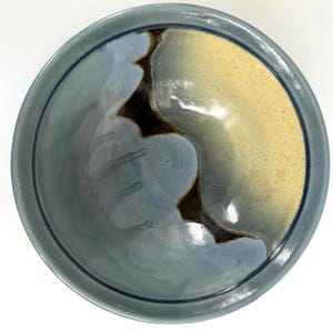 May include: A handmade ceramic bowl with a unique design. The bowl features a blend of colors, including blue, tan, and brown, creating an abstract pattern. The rim is a darker blue, and the interior has a glossy finish. This decorative bowl is perfect for serving or display.