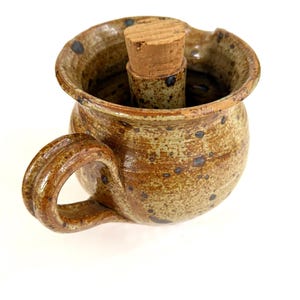 May include: A handmade ceramic mug with a handle, featuring a cork stopper. The mug is a speckled brown with dark blue spots and a rustic, textured finish. The cork stopper is cylindrical and fits snugly inside the mug's opening.
