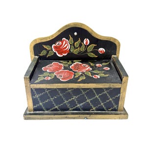 May include: A black wooden wall-mounted box with a hinged lid, featuring hand-painted red roses with green leaves on the top and back panel. The front panel displays a gold-painted wavy grid pattern, and the edges are trimmed in gold paint.