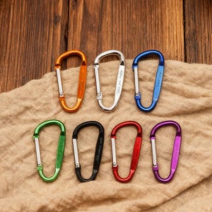 Könnte beinhalten: Sieben farbenfrohe Karabinerhaken in Orange, Silber, Blau, Grün, Schwarz, Rot und Lila. Sie sind auf einem beigen Stoff mit Holzgrund angeordnet. Einige Karabiner haben Text. Diese Karabiner sind ein nützliches Zubehör.