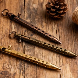 Könnte beinhalten: Drei Metallstifte in Bronze-, Messing- und Kupfertönen liegen auf einer Holzoberfläche. Die Stifte haben einen Ring oben und eine Reihe von Löchern entlang des Schafts. Ein Stift ist mit dem Namen "DAVID" graviert, ein anderer mit "JAMES".