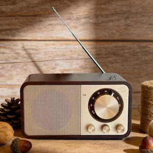 May include: A vintage-style radio with a brown wooden frame and a gold-colored front panel. The radio has a tuning dial, control knobs, and a silver antenna. The initials "M S" are visible on the top. The radio is on a wooden surface.