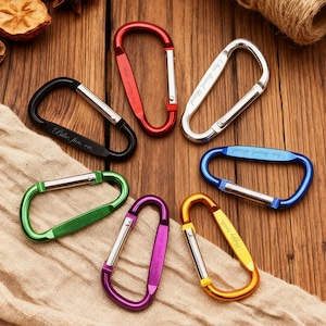 Könnte beinhalten: Eine Sammlung von sieben farbigen Karabinerhaken in Schwarz, Rot, Silber, Blau, Grün, Lila und Gold. Jeder Haken hat einen silbernen Stab und Text. Die Karabinerhaken werden auf einer Holzoberfläche und einem beigen Tuch präsentiert.