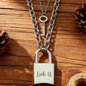 May include: A silver-colored necklace set featuring a padlock pendant engraved with "Lock U", a key pendant, and a chunky chain. The pendants hang on a wooden surface with pine cones and twine.