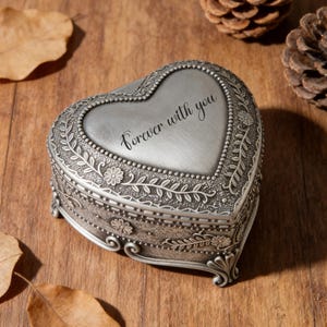 Könnte beinhalten: Eine silberfarbene herzförmige Schmuckschatulle mit der Aufschrift "Forever with you". Die Schatulle hat florale und Rankenverzierungen an den Rändern. Sie liegt auf einer Holzoberfläche mit getrockneten Blättern und Tannenzapfen.