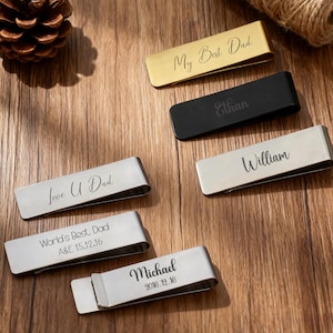Könnte beinhalten: Eine Sammlung personalisierter Geldklammern in Gold, Schwarz und Silber. Jede Klammer hat eingravierten Text, darunter Namen und Sätze wie "My Best Dad", "Ethan" und "Love U Dad". Die Klammern werden auf einer Holzoberfläche präsentiert.