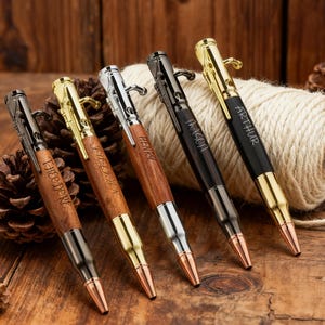 Könnte beinhalten: Fünf personalisierte Stifte mit kugelförmigen Spitzen. Die Stifte haben Holz- und schwarze Schäfte mit goldenen, silbernen und schwarzen Akzenten. Jeder Stift ist mit einem anderen Namen graviert: Theodore, Henry, Jason und Arthur. Die Stifte werden auf einer Holzoberfläche präsentiert.