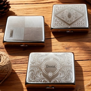 May include: Three silver metal cigarette cases with engraved designs. One case has a geometric pattern with the name "William", another has a diamond pattern with the name "Alan", and the third has a floral design with the name "David".