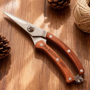 Könnte beinhalten: Gartenschere aus Edelstahl mit Holzgriffen. Die Schere hat eine scharfe, spitze Klinge und den Text "Best Dad Ever" auf einem Griff eingraviert. Die Schere liegt auf einer Holzoberfläche mit Tannenzapfen und Bindfaden.