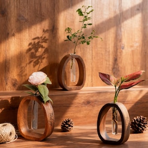 Könnte beinhalten: Drei Holzvasen mit Glasröhren, jede mit einer Pflanze oder Blume. Die Vasen haben ein ovales Design und sind aus dunklem Holz gefertigt. Eine Vase hat eine rosa Blume, eine andere grüne Blätter und die dritte rote und grüne Blätter. Eine Schnurkugel und Tannenzapfen sind ebenfalls vorhanden.