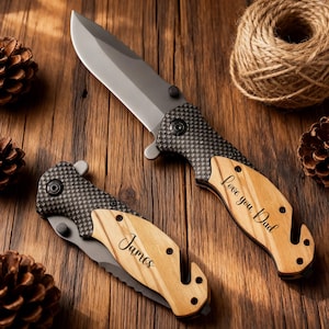 Könnte beinhalten: Zwei Klappmesser mit silbernen Klingen und Holzgriffen werden auf einer Holzoberfläche gezeigt. Eines ist mit "Love you, Dad" graviert, das andere mit "James". Die Griffe haben ein Karbonfasermuster in der Nähe der Klinge.