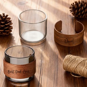 Könnte beinhalten: Klares Glas mit einer Holzmanschette mit der Gravur "Best Dad Ever". Ein Holzarmreif ist mit "Daniel" graviert. Die Artikel befinden sich auf einer Holzoberfläche mit Tannenzapfen und Bindfaden.