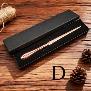 May include: A copper-colored letter opener with the word "Henry" engraved on the handle, presented in a black box. The opener is displayed on a wooden surface, accompanied by a pine cone and a spool of twine.