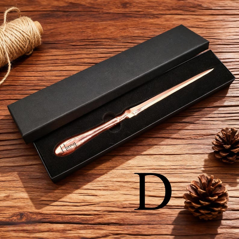 May include: A copper-colored letter opener with the word "Henry" engraved on the handle, presented in a black box. The opener is displayed on a wooden surface, accompanied by a pine cone and a spool of twine.
