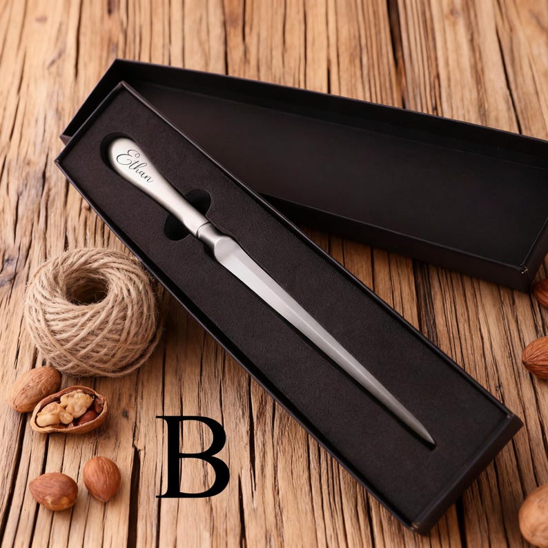 May include: A silver letter opener with the name "Ethan" engraved on the handle, presented in a black gift box. The opener has a long, pointed blade. The box is open, revealing the opener against a black interior. The background is a wooden surface.