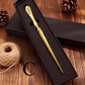 May include: A gold-colored letter opener with the name "William" engraved on the handle, presented in a black gift box. The opener has a long, pointed blade. The background includes a wooden surface, a ball of twine, and pine cones.