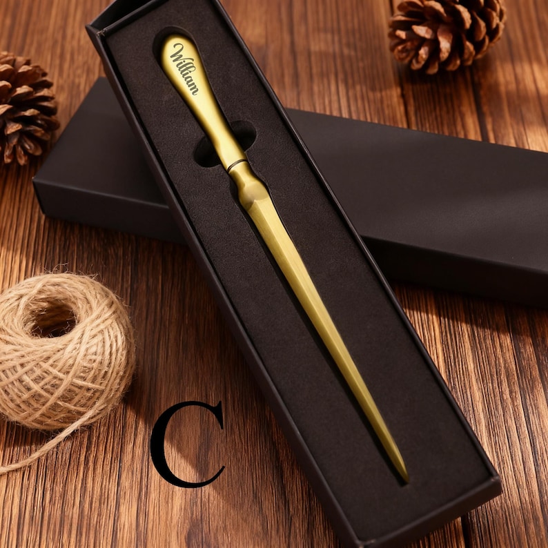 May include: A gold-colored letter opener with the name "William" engraved on the handle, presented in a black gift box. The opener has a long, pointed blade. The background includes a wooden surface, a ball of twine, and pine cones.