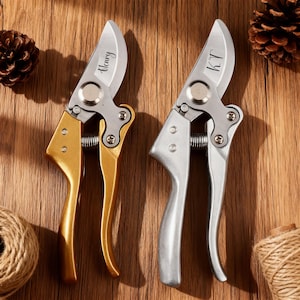 Könnte beinhalten: Zwei personalisierte Gartenscheren mit silbernen Klingen und goldenen und silbernen Griffen. Auf einer Schere steht "Henry", auf der anderen "K.T.". Die Scheren liegen auf einer Holzoberfläche, mit Tannenzapfen und Bindfaden.