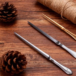 May include: Three letter openers on a wooden surface. Two are silver-toned, one with the name "James" engraved. The third is gold-toned. A pinecone and a roll of twine are also present.