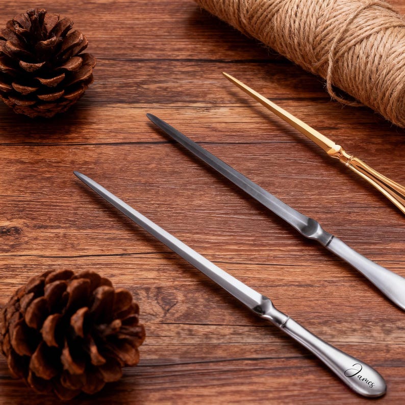 May include: Three letter openers on a wooden surface. Two are silver-toned, one with the name "James" engraved. The third is gold-toned. A pinecone and a roll of twine are also present.
