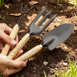 Peut inclure: Un ensemble de trois outils de jardinage avec des manches en bois et des têtes en métal noir. Les manches portent les inscriptions "To the best dad ever" et "Happy Birthday". L'ensemble comprend une petite pelle, une fourche à trois dents et un petit râteau.