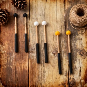 May include: Six percussion mallets with wooden handles and black grips. The mallet heads are black, white, and yellow. The mallets are arranged on a rustic wooden surface with pine cones and twine.