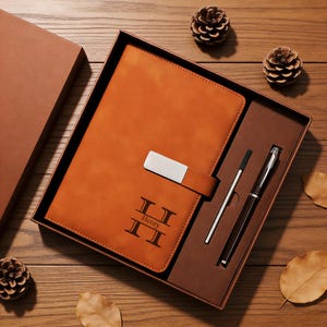 Könnte beinhalten: Ein braunes Leder-Notizbuch- und Stift-Set in einer Geschenkbox. Das Notizbuch hat einen silbernen Verschluss und den Namen "Henry" auf dem Einband eingraviert. Ein silberner Stift und ein schwarzer Stift sind enthalten.