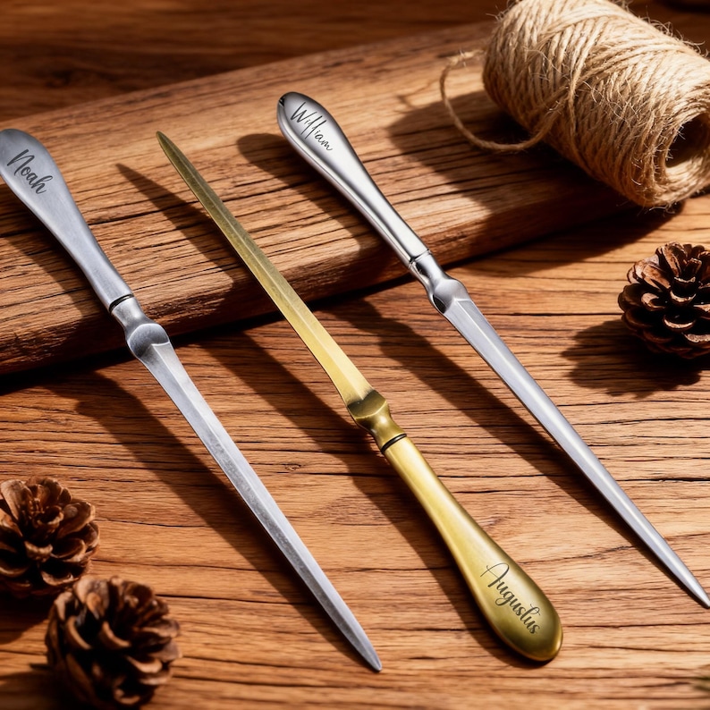 May include: Three letter openers with personalized names: "Noah", "William", and "Augustus". The openers are silver and gold-toned, with a pointed blade and a handle. The items are arranged on a wooden surface.