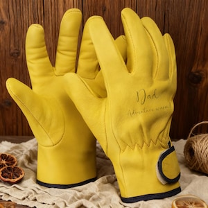 May include: Yellow leather work gloves, featuring the word "Dad" and the phrase "Adventure warm!" on the right glove. The gloves have dark blue trim. They are displayed on a beige cloth.