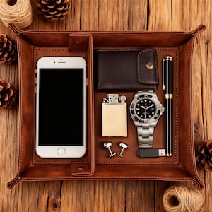 May include: A brown leather valet tray with a smartphone, wallet, lighter, watch, pen, cufflinks, and USB drive. The tray is divided into compartments and sits on a wooden surface. Decorative pinecones and twine are also present.