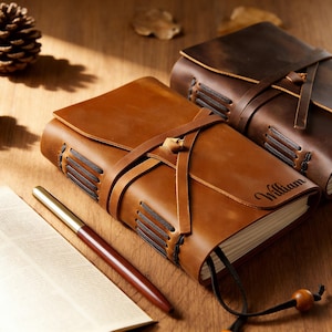 Könnte beinhalten: Zwei Lederjournale mit Riemenverschlüssen werden auf einer Holzoberfläche gezeigt. Das vordere Journal ist hellbraun und trägt den Namen "William" auf dem Einband. Ein Stift und ein offenes Buch befinden sich im Vordergrund. Das zweite Journal ist dunkelbraun.