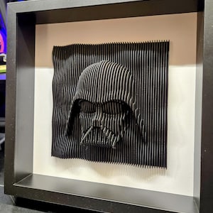 Puede incluir: Un casco de Darth Vader impreso en 3D en blanco y negro en un marco negro. El casco está hecho de papel en capas y tiene una superficie texturizada.